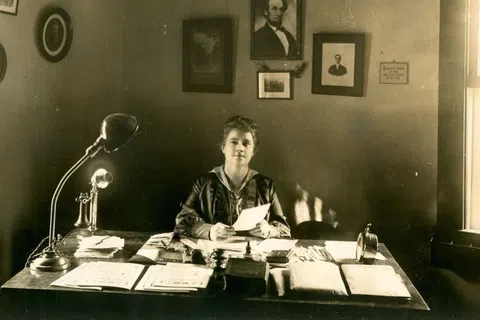 Early 20th-century woman sitting at desk with papers and vintage telephone under portraits on wall
