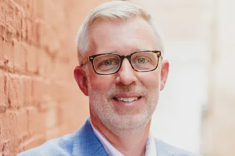 Smiling man with gray hair and glasses wearing a blue blazer stands against a brick wall outdoors.