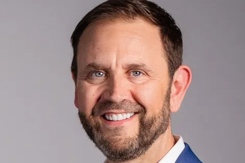 Smiling man with beard wearing a blue blazer and white shirt against a plain gray background