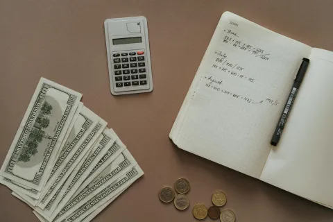 Flat lay of cash, calculator, notebook with handwritten notes, pen, and assorted coins on a brown surface