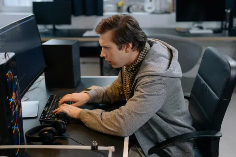 Young man wearing a gray hoodie working intensely on a desktop computer in an office setting.
