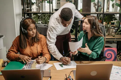 Diverse team discussing work with laptops and headsets in a modern office filled with plants and documents.