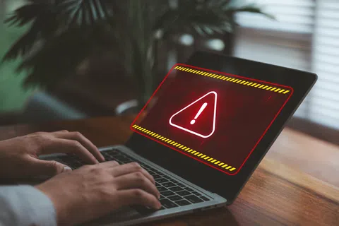 Person typing on laptop showing red warning alert with exclamation mark on screen in a home setting.