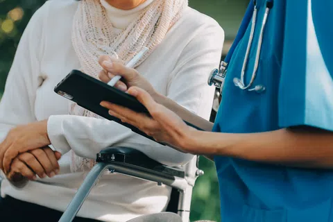Healthcare professional consulting an elderly patient in a wheelchair outdoors using a tablet device.