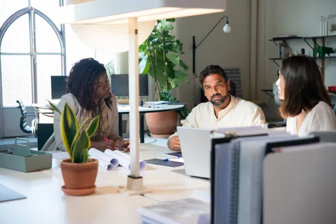 Three diverse coworkers engaged in a meeting around a white office table with laptops and documents in a bright workspace.