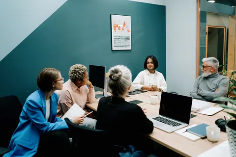 Diverse team of five people having a meeting around a wooden table in a modern office with teal wall.
