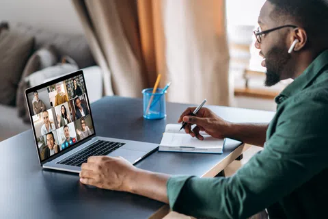 Man participating in a virtual video conference on laptop while taking notes in a notebook at home.