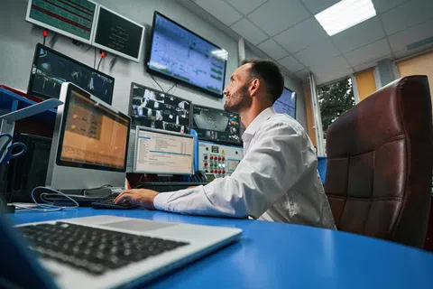 Man in white shirt monitoring multiple screens in a control room with security footage and data.