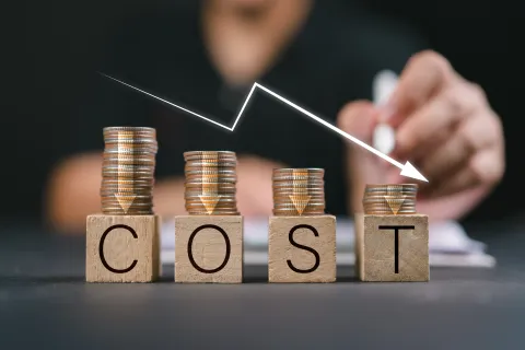 Stacked coins on wooden blocks spelling COST with a downward arrow indicating decreasing expenses.