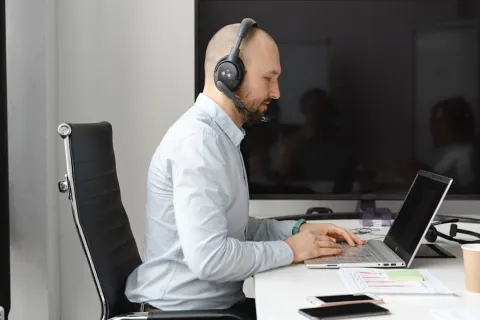 Man wearing headset working on laptop at office desk with smartphone and documents nearby