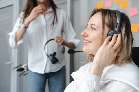 Smiling woman wearing headset in office with another person holding headset and sticky notes on wall behind.