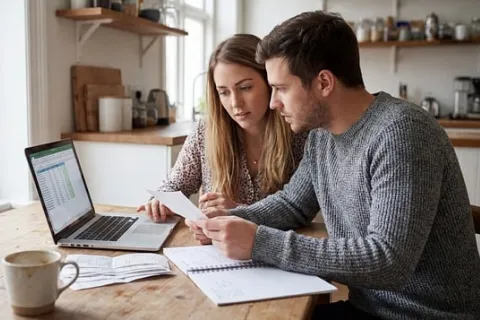 Young couple reviewing finances together at a wooden kitchen table with laptop, receipts, and notes.