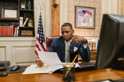 Man in suit reviewing documents at desk in office with American flag and computer monitor