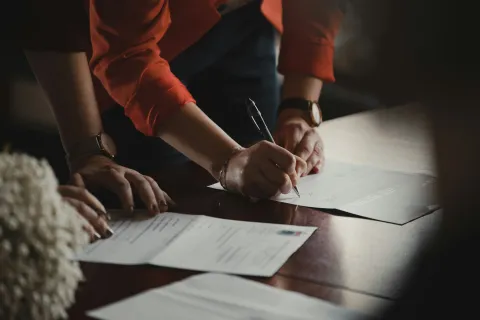 Person in red shirt signing documents on wooden table with papers in low light setting