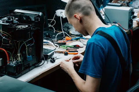 Young technician repairing electronic device at cluttered workbench with tools and wires.