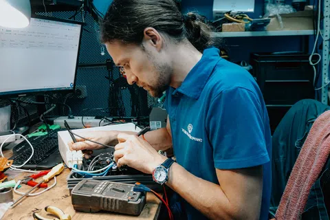 Technician in blue polo using multimeter to test electronic components on a cluttered workbench in workshop.