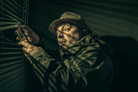 Man in a hat and jacket cautiously peeking through window blinds in a dimly lit room at night.