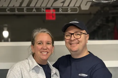 Smiling man and woman wearing matching cleaning service uniforms standing behind a counter indoors.