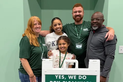 Five diverse people smiling behind a sign promoting Lapels dry cleaning and garment care services.