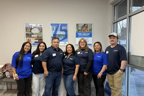 Group of seven smiling employees in branded shirts standing inside a service center with posters.