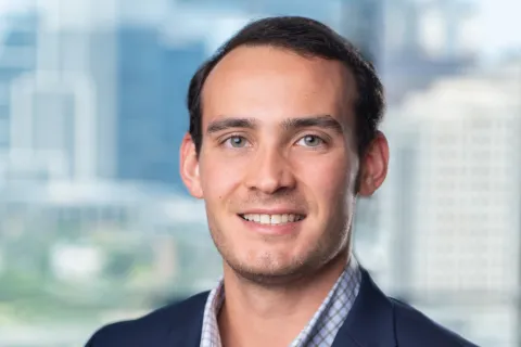 Young man smiling wearing a navy blazer and checked shirt with a cityscape background