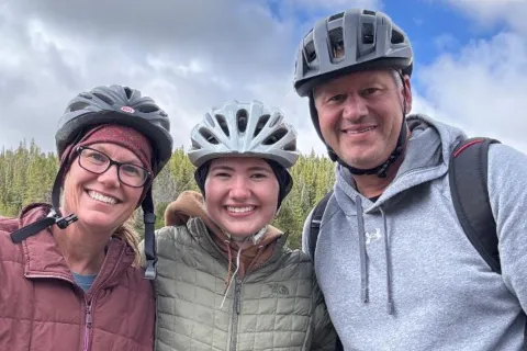 Three smiling people wearing bike helmets outdoors with forest and cloudy sky in background