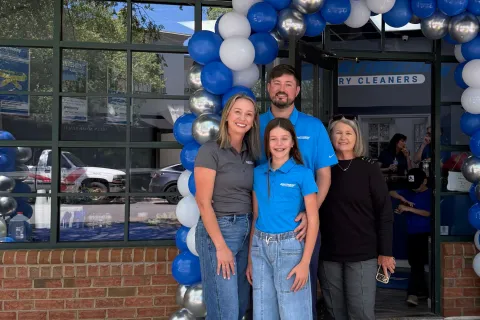 Happy family standing under a blue, white, and silver balloon arch in front of a dry cleaners storefront.