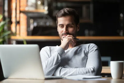 Man in a white sweater thoughtfully working on a laptop with a pen in hand in a cozy workspace.