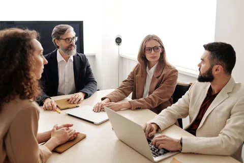 Four professionals engaged in a business meeting around a table with laptops and notebooks in a bright office.
