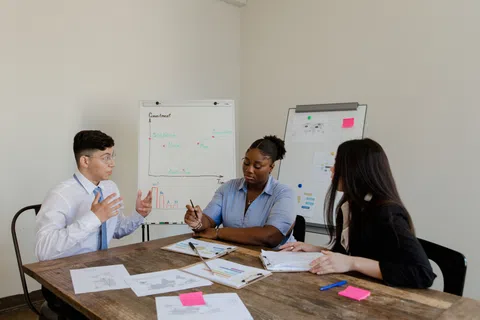 Three colleagues in a meeting discussing charts and graphs in a modern office setting with whiteboards.
