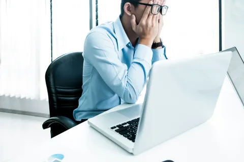 Stressed man in blue shirt with glasses sitting at desk covering face in front of laptop in real estate office in New Jersey.