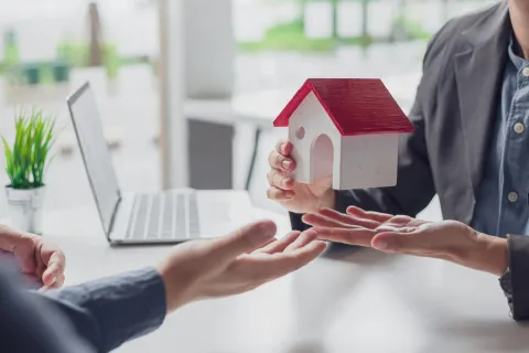 Real estate agent handing over a small house model to a client - Computer in background needing IT services