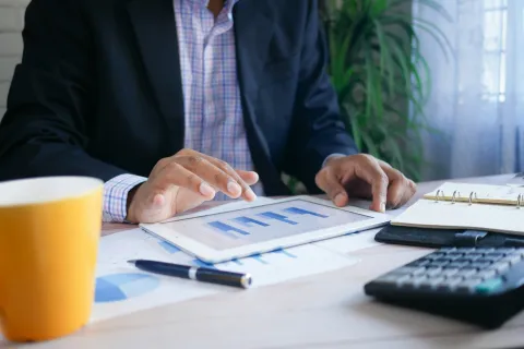 Person analyzing financial bar charts on a tablet with pen, calculator, notebook, and coffee cup on desk.
