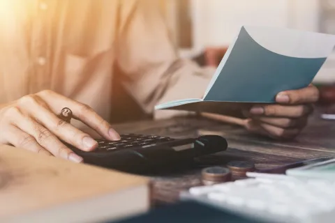 Person using calculator and holding checkbook at table trying to save money on IT expenses