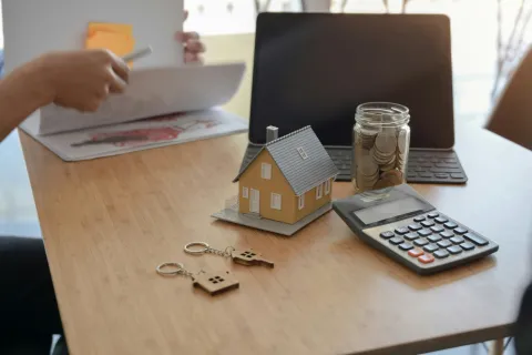 Desk with house model, calculator, coin jar, house keychains, and person reviewing documents near laptop for real estate firms in New Jersey.