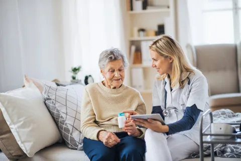 Senior woman receiving medical care from a female home healthcare provider holding a pill bottle and a tablet.