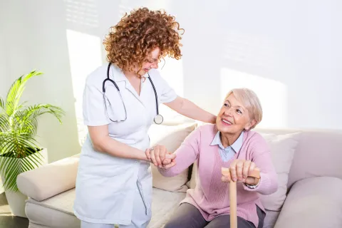 Young female nurse assisting elderly woman stand with walking stick in a bright room, showing home healthcare services in new jersey