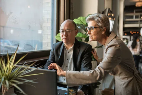 Two professionals collaborate on a laptop near a window in a modern office setting with plants and coffee.
