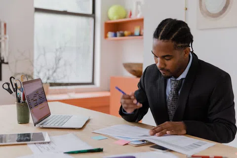 Professional man in suit reviewing documents at desk with laptop and office supplies near bright window.