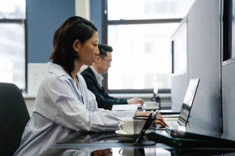 Two professionals working on laptops in a modern office with large windows and cubicles.
