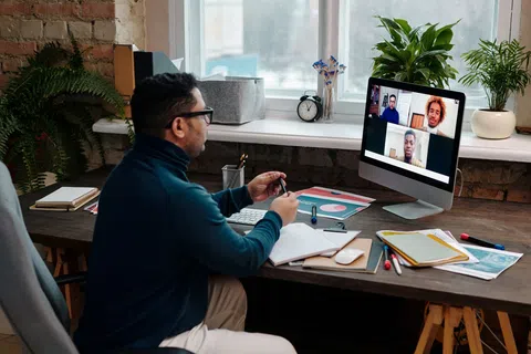 Man attending a virtual meeting on desktop computer in a home office with notebooks and plants around.