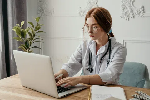 Female doctor with stethoscope working on laptop at desk with notebook and pills nearby