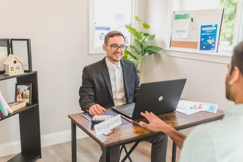 Mortgage broker consulting a client in an office with laptop, calculator, and financial documents on the desk
