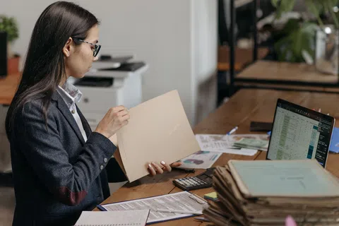 Businesswoman reviewing documents at desk with laptop, calculator, and paperwork in an office setting.