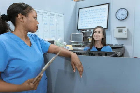 Two medical professionals in blue scrubs talking in a hospital nurse station with computer and whiteboard.