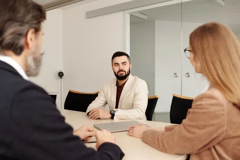 Man in beige blazer sitting at conference table engaged in discussion with two colleagues in office setting.