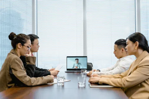 Four people in a meeting room engaged in a video conference with a person on a laptop screen.