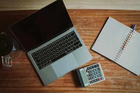 Laptop, calculator, spiral notebook, pen, and glass mug on wooden desk workspace