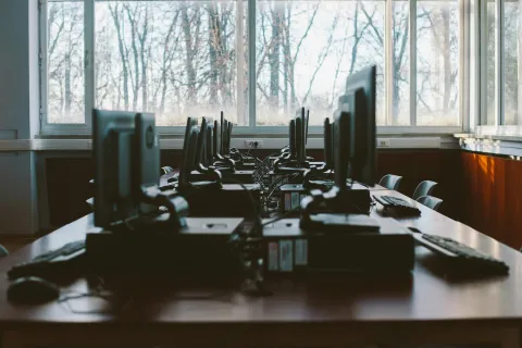 Empty computer lab with rows of monitors and keyboards on wooden desks by large windows with winter trees outside