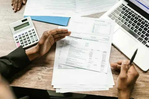 Person reviewing bank statements with calculator and laptop on wooden desk for finances.